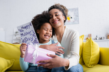 pleased african american woman holding gift box and embracing daughter with happy mothers day card