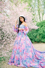 Fototapeta premium Beautiful young asian brunette woman in luxury long dress among the blossoming sakura flowers. enjoying spring Early spring concept. Rest on nature vacation. Girl in straw hat.