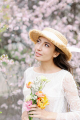 Beautiful young caucasian brunette woman in luxury long dress among the blossoming sakura flowers. enjoying spring Early spring concept. Rest on nature vacation. Girl in straw hat.