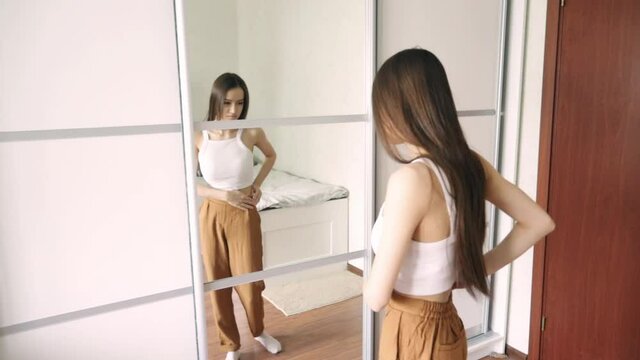 the cult of a beautiful body, public opinion, imposed stereotypes, A teenage girl is looking for fat on her body, stands at home in front of a mirror