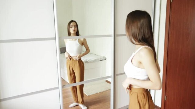 slender teen girl looking for fat on her body, standing at home in front of the mirror