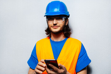 Studio portrait of young thoughtful construction worker engineer wearing safety equipment using smartphone on the background of grey wall with copy space.