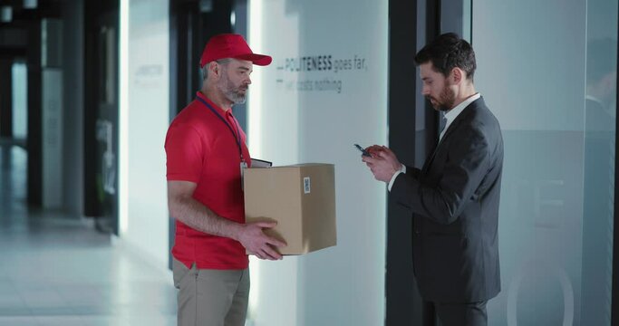 Fast delivery. Courier in red uniform giving carton box to the customer. Young adult businessman paying with smartphone receiving parcel at the office.