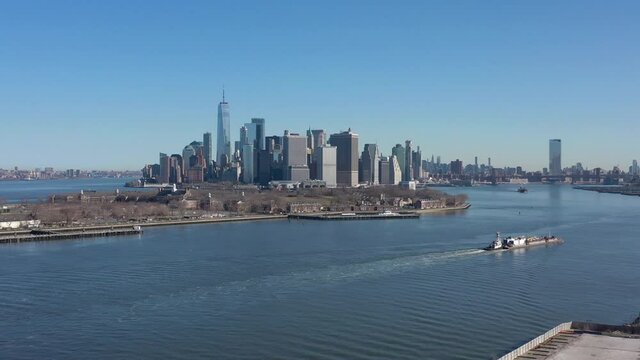 An aerial view of New York harbor on a sunny day with clear blue skies. The camera boom up high enough to see Governor's Island, lower Manhattan in the background and two barges.
