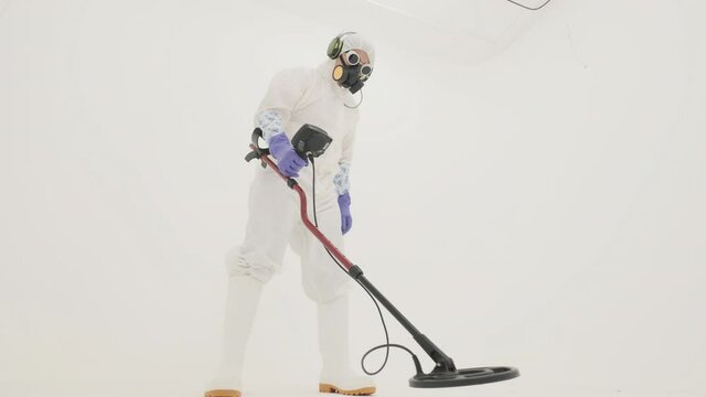 Man in white chemical protection suit, gas mask and purple rubber gloves holds metal detector and searches for something around. Filmed on white background.
