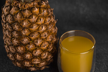 Pineapple fruit and juice in double glass cup on black stone background. Pouring yellow tropical fruit juice into glass.