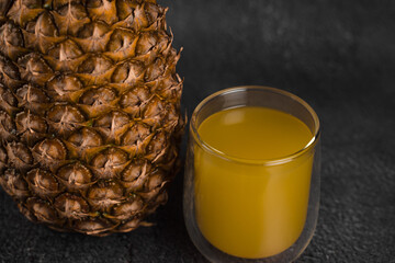 Pineapple fruit and juice in double glass cup on black stone background. Pouring yellow tropical fruit juice into glass.