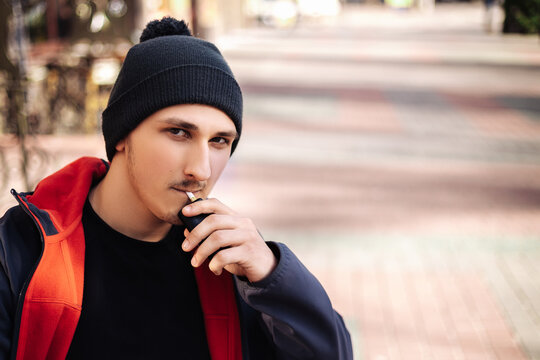 Young Adult Man Outside And Smoking Tobacco Device Electronic Cigarette Heater. Smoke And Steam System With Sticks Inside, Image With Copy Space. Harmful Habit Harm To Health Lungs
