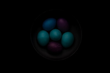 Blue, turquoise, and purple easter eggs in a bowl with strong shadows and dark background.