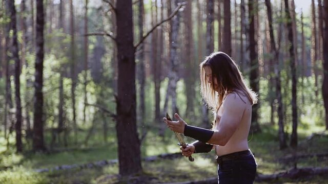 In the middle of the forest stands a man with a bare torso and holds a large knife in his hands. The light from the sun reflects off the weapon and falls on the face