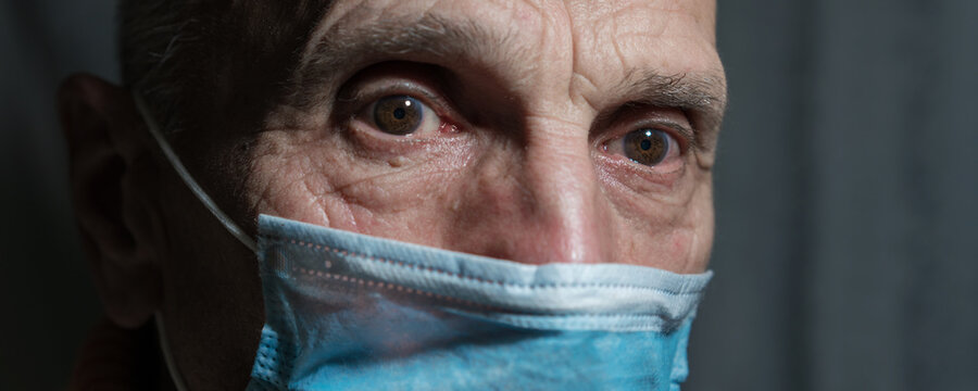 Face Of Adult Man Wearing Medical Facemask Stands In Dark Hospital Room.