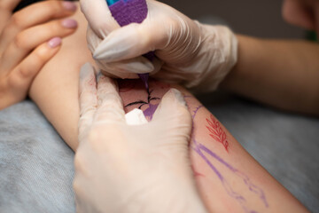 Master tattoo draws the red paint on the clients tattoo. Tattoo artist holding a pink tattoo machine in black sterile gloves and working on the professional blue mat.