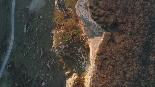 Top view of rocky cliff line. Shot. Rocky cliff is illuminated by sun's rays on background of dark plain at foot. Beautiful rocky cliff in morning or evening sun