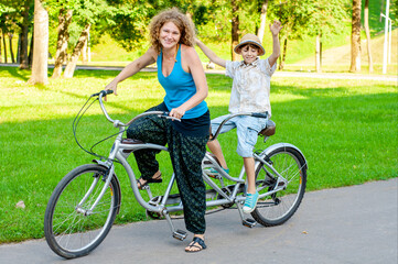 Trendy mom and son schoolboy ride together in a summer park on a bicycle tandem. Family healthy...