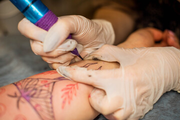 Master tattoo draws the red paint on the clients tattoo. Tattoo artist holding a pink tattoo machine in black sterile gloves and working on the professional blue mat.