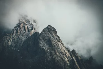 Poster Donker sfeervol surrealistisch landschap met donkere rotsachtige bergtop in lage wolken in grijze bewolkte hemel. Grijze lage wolk op hoge top. Hoge zwarte rots met sneeuw in lage wolken. Surrealistische sombere bergen. © Daniil