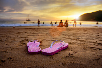 Beach sandal or pink flip flops for women on sandy beach with tourist group playing water waves crashing on the beach in beautiful dramatic sunset background and flare.