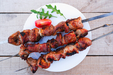 Juicy meat on skewers with tomatoes and herbs on a white plate on a wooden background. Kebab of meat is fried on the grill.