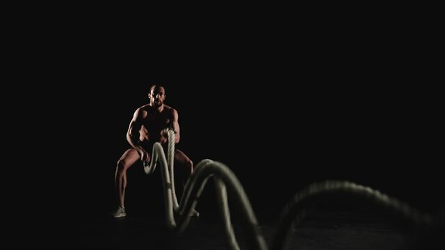 Athletic man training with battle ropes on black background. Slow motion