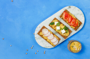 Vegetarian appetizer, rye bread with hummus, slices of fresh vegetables, herbs and tofu. On a bright blue background, overhead