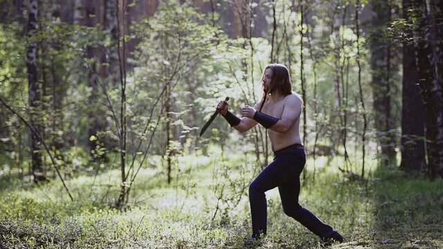 In a sunny clearing in the forest, a man is training with a blade in his hands. Cool slow-motion shots. The guy's torso is bare