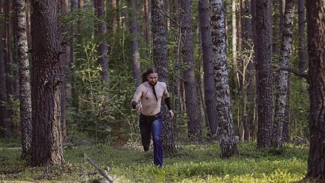 A man with a knife and a bare torso runs through the forest among the trees. He has a beard. Shooting in slow motion