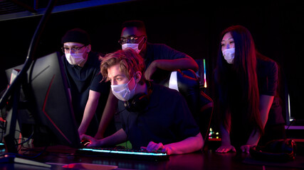 Two guys and a girl in medical masks in a computer club give support to their friend during his game and rejoice