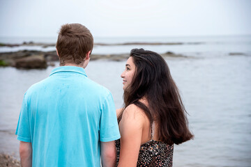 Casual outdoor back view portrait of a cute young couple in love walking close together on the beach with the woman's face in profile as she looks at the man  
