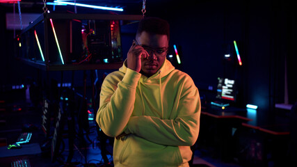 A brooding black guy in a hoodie puts on his glasses in the neon lights of the computer room