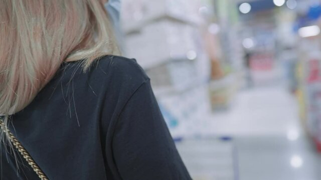 View from behind of blonde woman wearing medical face mask pushing grocery shopping cart in stay safe in public during pandemic, over shoulder shot, shopping capitalist demand and supply, stock up goo