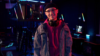 In a computer club in neon light, a young guy in a hat, glasses and a denim jacket poses for the camera and smiles
