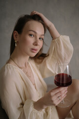 Young beautiful woman wearinng white vintage dress enjoying glass of red wine sitting in a chair near textured wall.