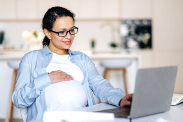 Concentrated joyful pregnant mixed race woman wearing glasses, sitting at table, uses laptop for...