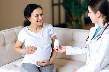 The doctor prescribes vitamins for a pregnant mixed race patient, sitting on the couch at home or in the hospital, the expectant mother stroking her belly. Healthy pregnancy concept