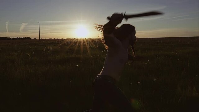 A man waves a blade in the evening in a field at sunset. He's practicing his punches