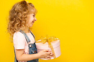 Little blonde girl on a yellow background holds a gift in a pink round box with gold ribbon, copy space