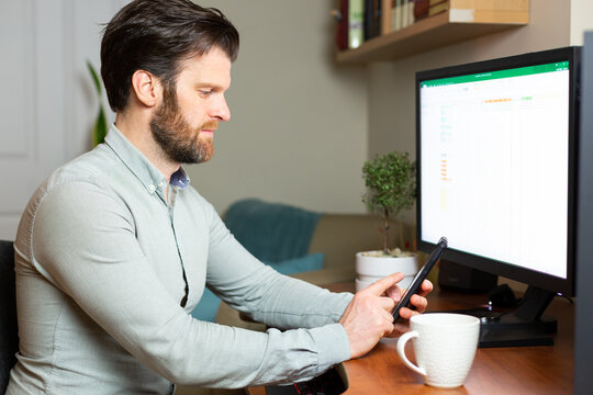 Man Taking Video Call On His Cell Phone Working From Home On Desktop Computer.