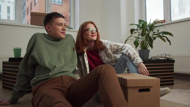 Couple sits on a parquet floor and examines the walls of a new apartment