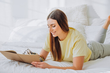 Smiling blonde woman in yellow t-shirt writes in paper notebook with metal bionic arm lying on bed in room at home closeup