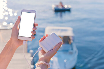 Mock-up of a smartphone in the hands of a girl on the pier. Charge your phone with Power Bank. Against the background of a river, lake, bay, with boats and a yacht.