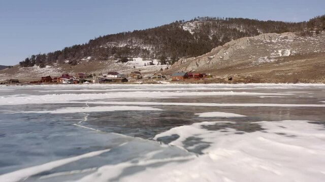 Aerial view, the drone flies forward at high speed close to the ice overlooking the village and rocky coastline. Uzury village with a coastline of rocks and hills covered with snow. Frozen Lake Baikal
