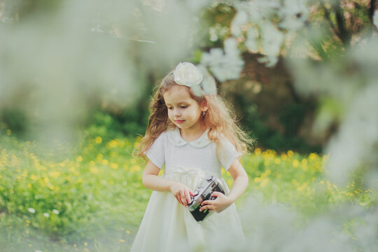 Little girl in white dress holding film camera in spring cherry garden. Portrait of happy child among white flowers trees. Childhood. Young photographer takes his first photos in sunny blooming park.