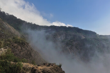 The rocky slopes of the mountains hide in the fog. Some green vegetation at the top. Blue sky with clouds. Vietnam. Dalat
