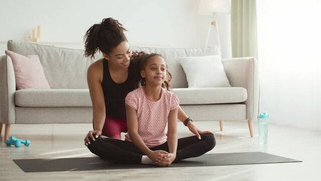 Cute african american girl sitting in lotus position on floor and her mother helping to stretch her legs