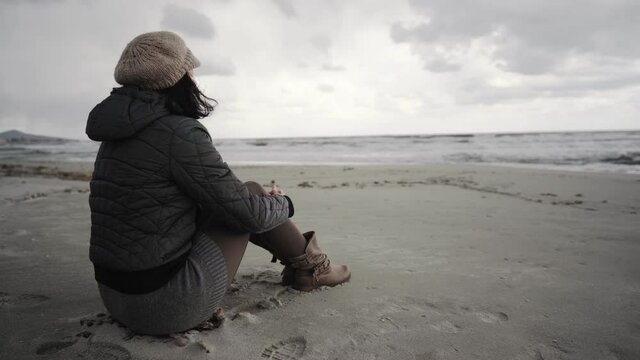 Sad and pensive lonely woman with winter clothes and woolen hat filmed from back sitting at the beach looks at the horizon over the ocean sea on a stormy day with dark clouds. Social isolation issue