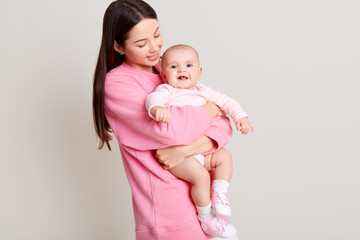 Dark haired young mother hugging her daughter, baby smiling, mommy looking at infant, wearing casual pink sweater and white pants, isolated over light background.