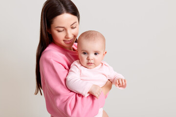Dark haired Caucasian mother with long dark hair holding her cute baby girl wearing bodysuit, woman in casual outfit looking at her daughter with love, isolated over white background.