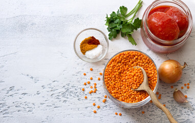 Ingredients for vegan tomato lentil sauce, soup or for lentil pancakes: red lentils, onions, garlic, canned tomatoes, spices and herbs on a light gray textured background, top view, space