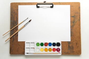 Art easel (drawing board) with blank paper, paint brushes and colourful of watercolour paint palette on white table background.