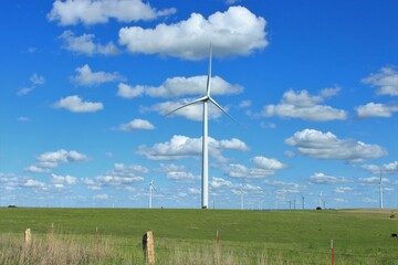 An Electric windmill in a farm pasture with green grass and a stone post fence with blue sky and...